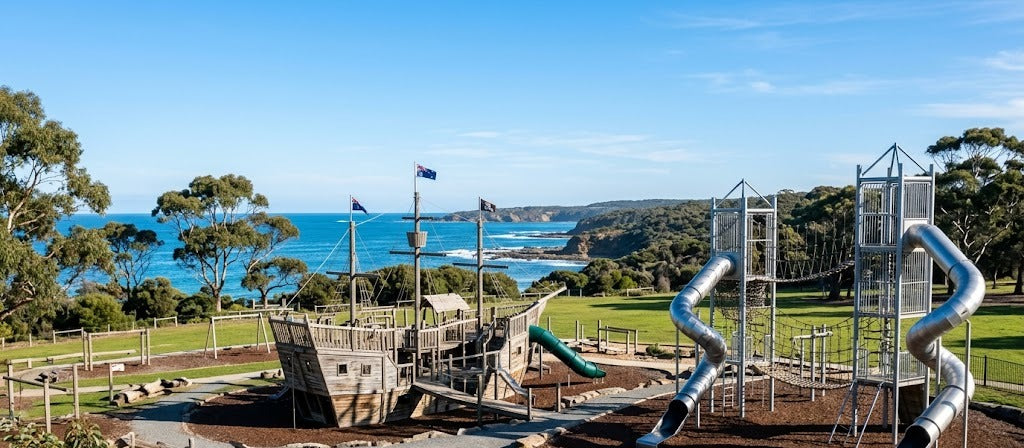 Large coastal adventure playground in Adelaide with a timber pirate ship, metal slides and climbing towers on green parkland by the sea